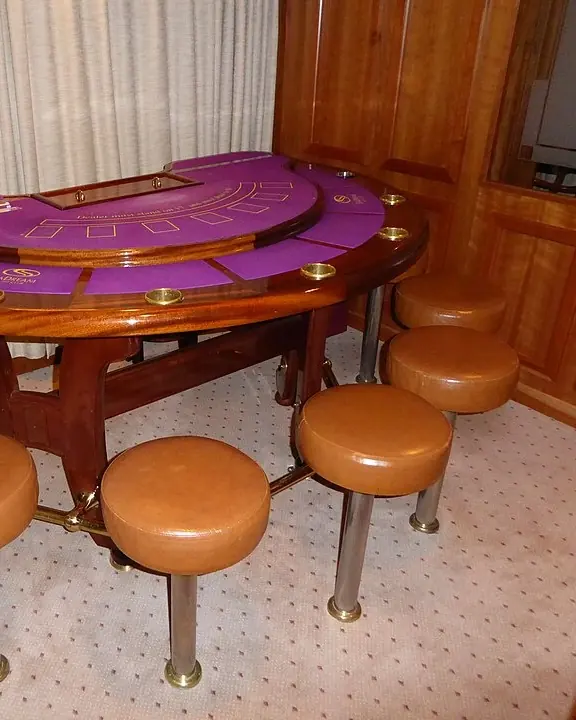 Félkörös blackjack-asztal lila posztóval, lakkozott fa kartámasszal és bőr ülőkés bárszékekkel egy hajó kaszinójában