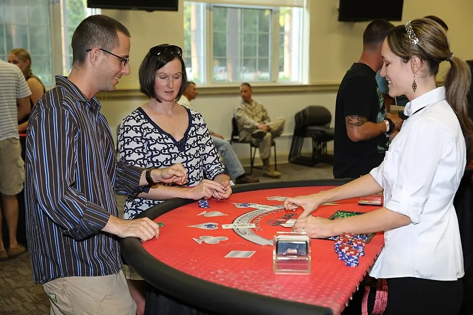 Osztó és két játékos egy piros posztós blackjack-asztalnál, az asztalon zsetonok és átlátszó kártyaadagoló